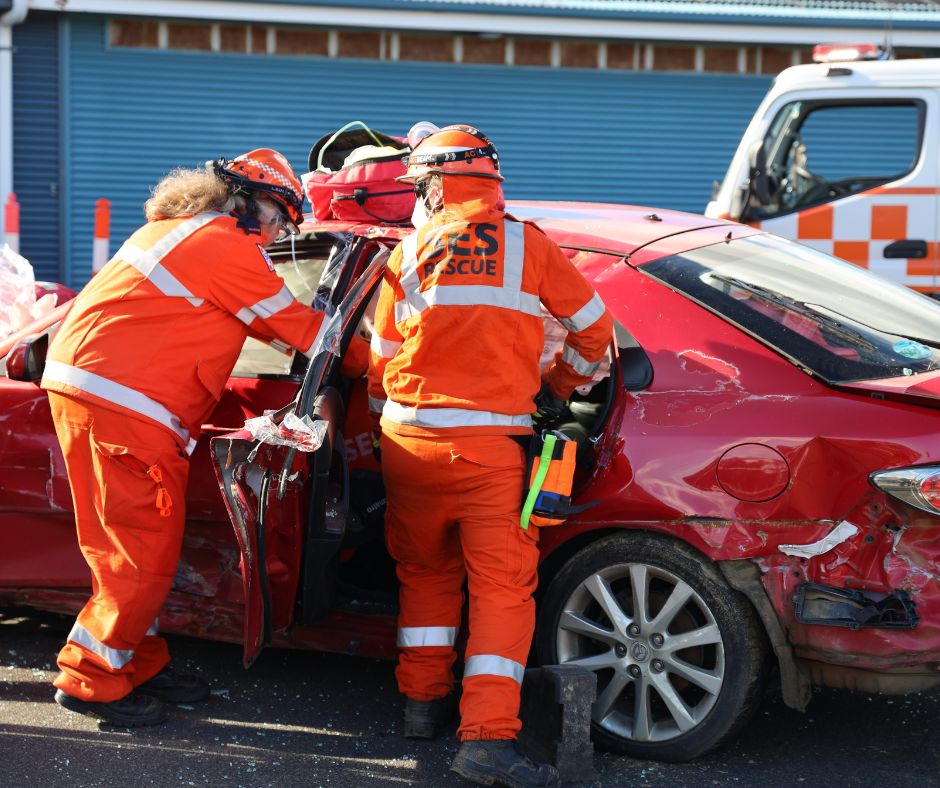 Two emergency rescue workers in orange uniforms attend to a damaged red car at the scene of an accident. The car’s side is heavily dented, and an emergency vehicle is visible in the background.