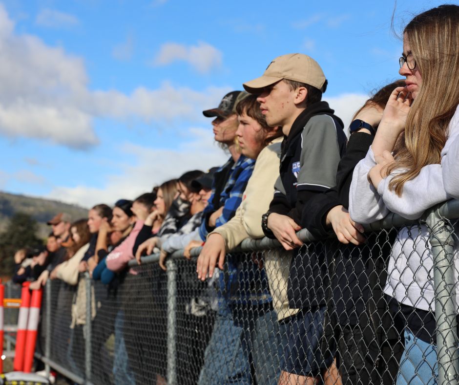 A group of people stand closely together behind a chain-link fence, watching something intently outdoors under a blue sky with scattered clouds.