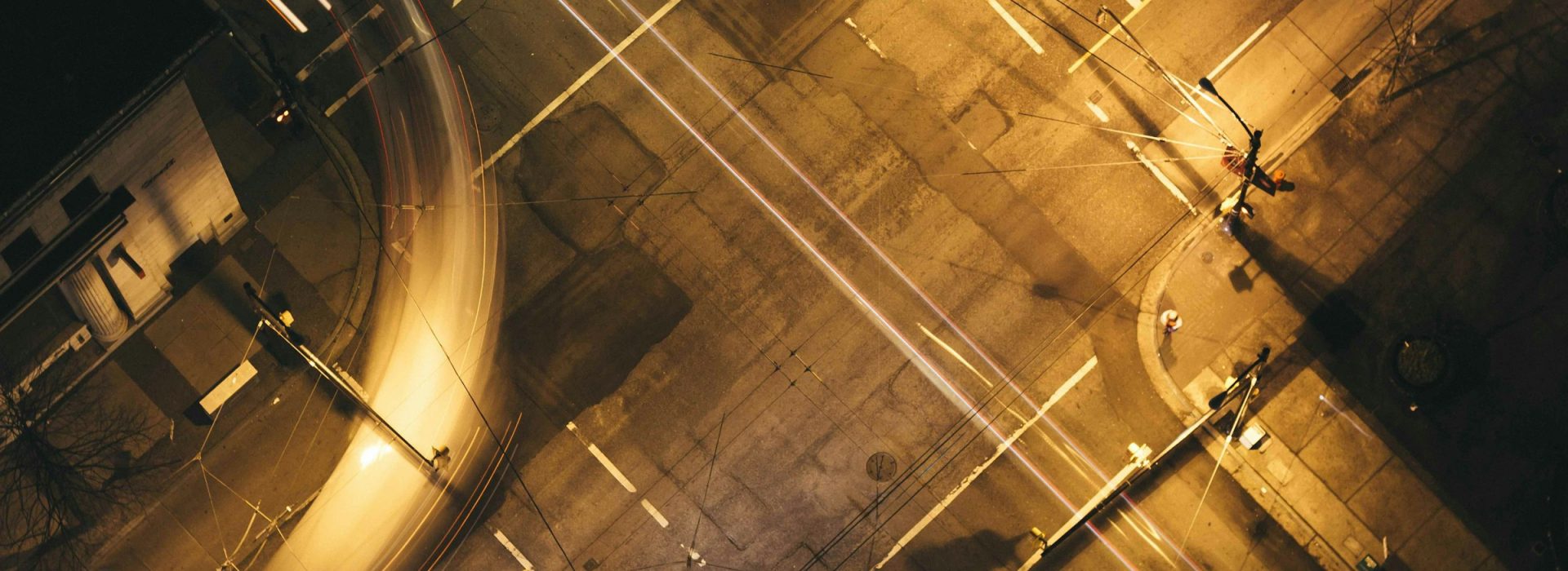 Aerial view of an empty city intersection at night, showing streaks of light from passing vehicles and illuminated traffic signals, with faint shadows and a few parked cars visible.