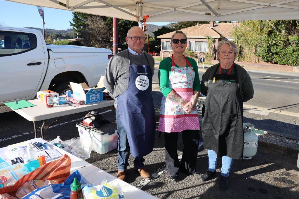 Three people stand under a market tent wearing aprons and smiling. Behind them is a table with food, supplies, and a white pickup truck parked by the roadside on a sunny day.