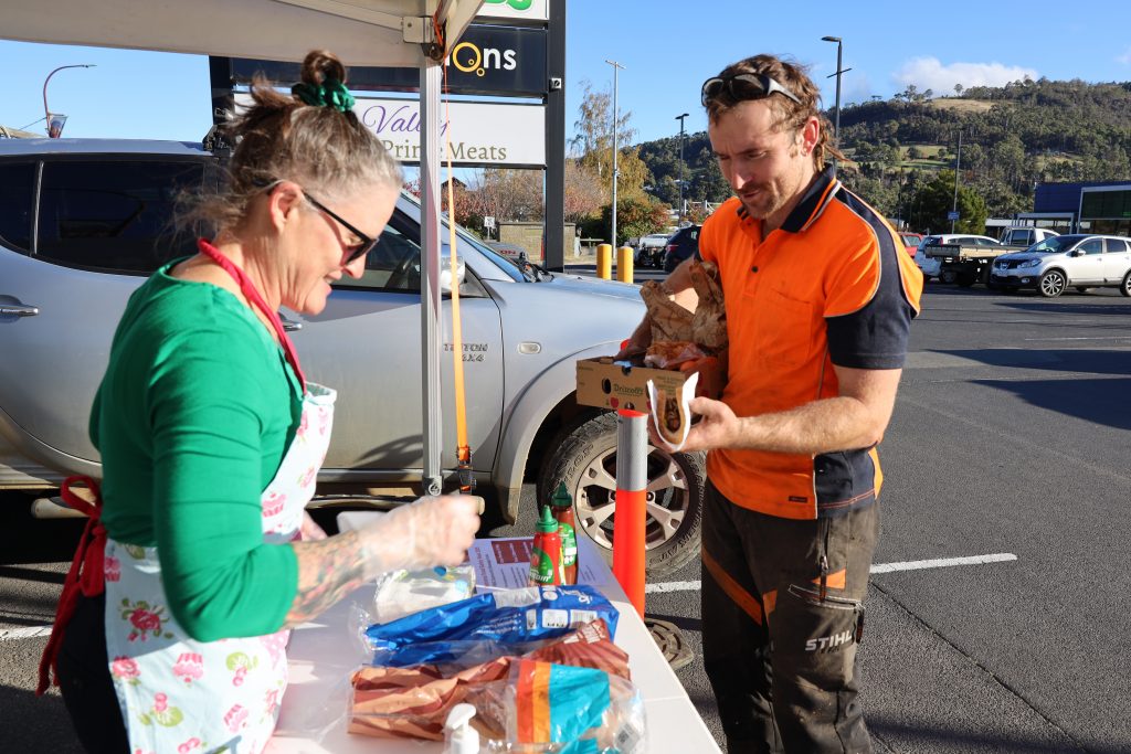 A woman wearing glasses and an apron serves food to a smiling man in an orange work shirt at an outdoor table in a parking lot, with a car and hills in the background.