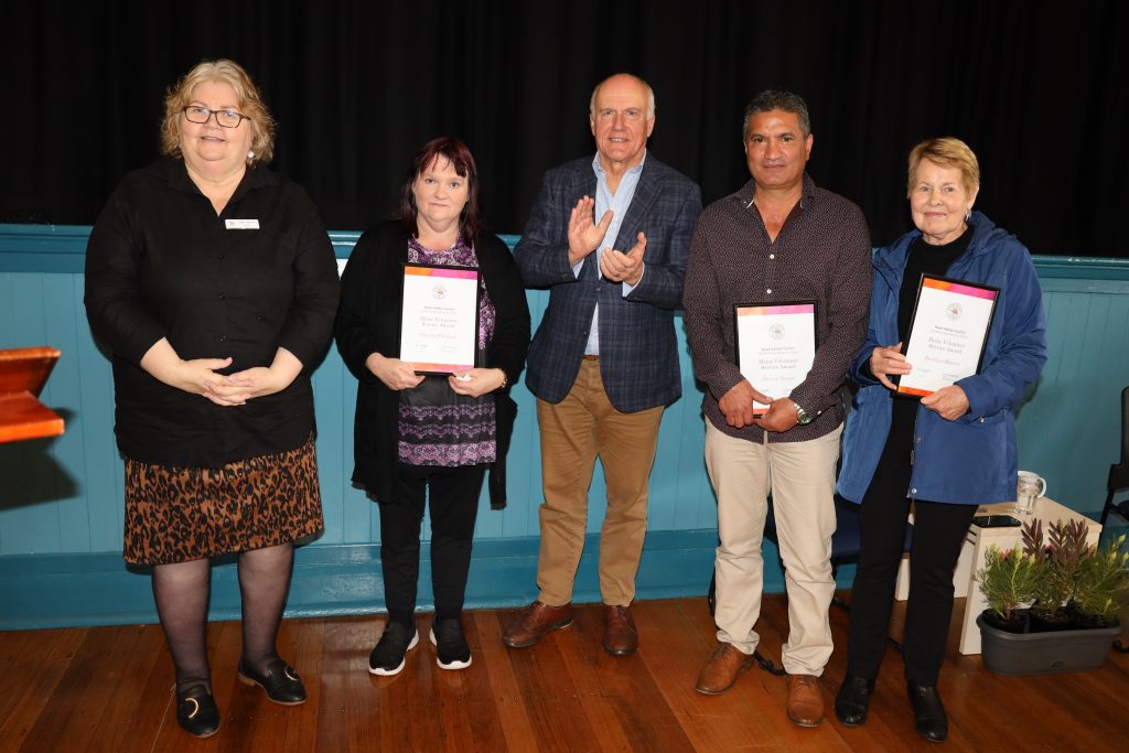 Five adults stand indoors on a wooden floor, four holding certificates. The group poses for a photo; one person is clapping. A dark curtain is behind them, and they appear to be at an award ceremony or presentation.