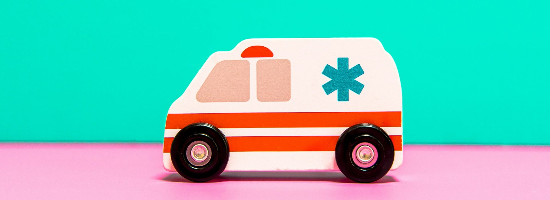 A small toy ambulance with black wheels, a red stripe, and a blue medical symbol is placed on a pink surface with a turquoise background.