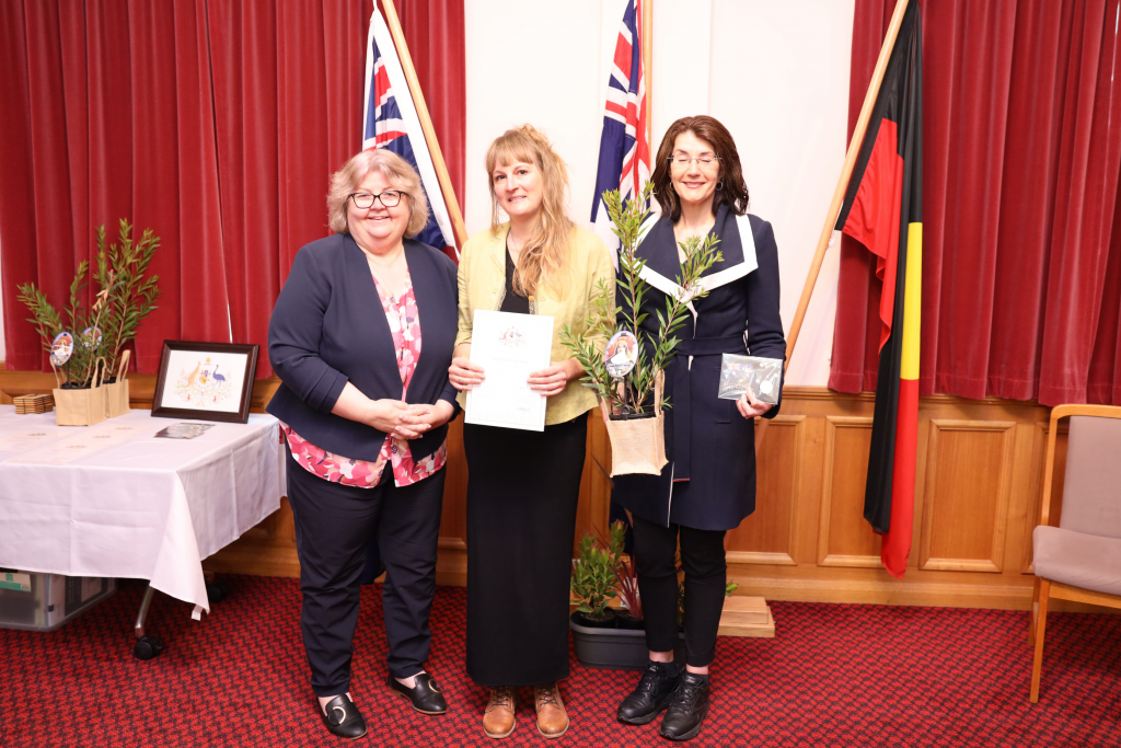 Three women stand indoors in front of Australian and Aboriginal flags. The woman in the center holds a certificate, and the woman on the right holds an olive tree. A table with documents and plants is visible on the left.