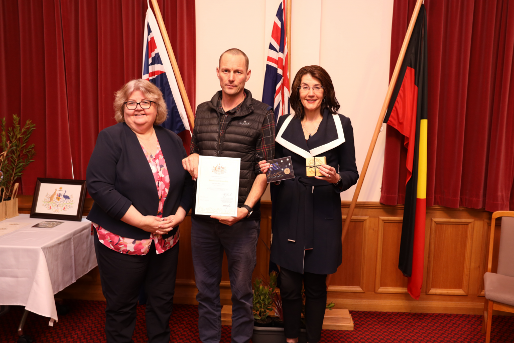 Three people stand indoors in front of Australian and Aboriginal flags. The man in the center holds a certificate; the woman on the right holds a box and medal. The woman on the left smiles. A table and framed art are in the background.