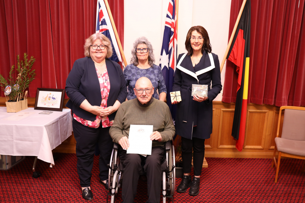 Four adults pose indoors for a group photo; three women stand behind a man in a wheelchair holding a certificate, with Australian and Aboriginal flags and a certificate table in the background.