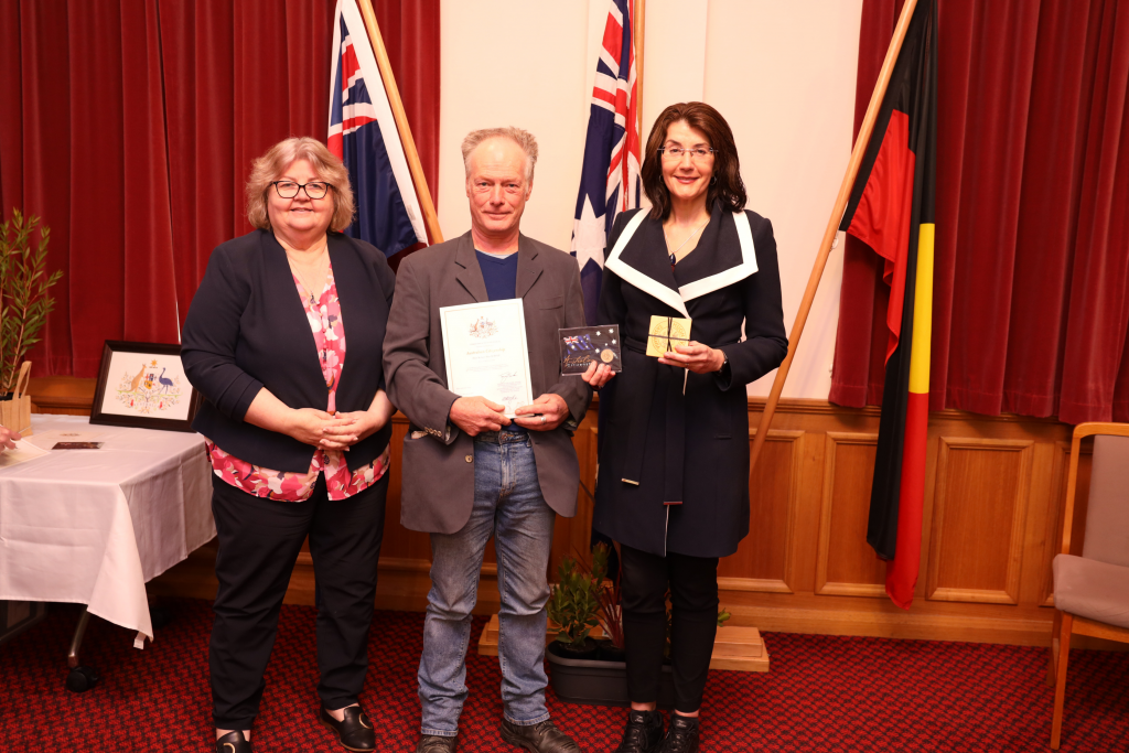 Three people stand indoors in front of Australian and Aboriginal flags, posing for a photo. The man in the center holds a certificate and booklet, while the women on either side smile at the camera.
