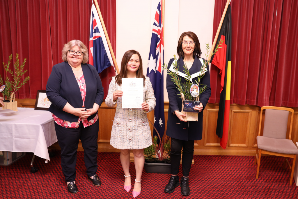 Three women stand indoors on a red carpet in front of three flags. The woman in the center holds a certificate, while the woman on the right holds a small plant in a basket. The woman on the left stands with her hands clasped.