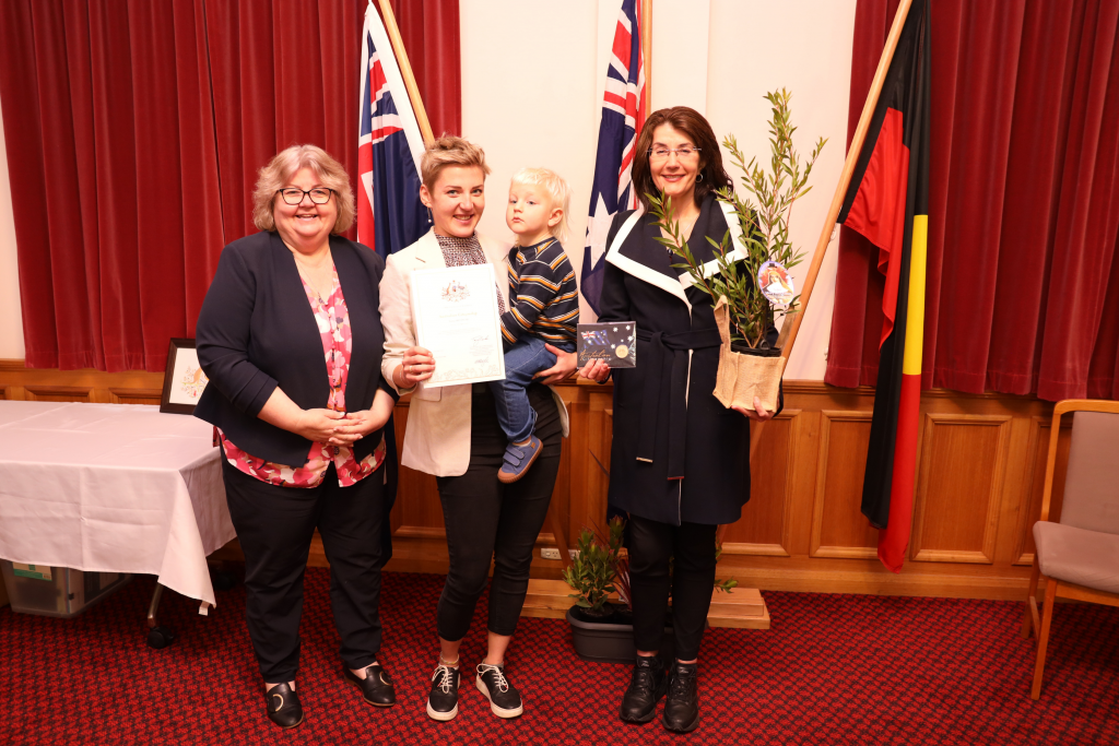 Four people stand indoors in front of Australian and Aboriginal flags. One holds a certificate, another holds a child, and a third person holds a potted plant. All are smiling for the photo.