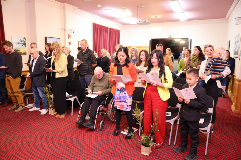 A group of people, including men, women, a child, and a person in a wheelchair, stand in a room holding song sheets and singing. The room has red carpet, framed pictures, and potted plants in the foreground.