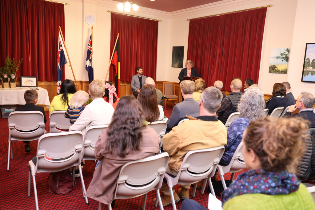 A group of people seated facing a woman speaking at a podium in a room with red curtains, Australian flags, and framed pictures on the walls. Two men are seated next to the speaker.