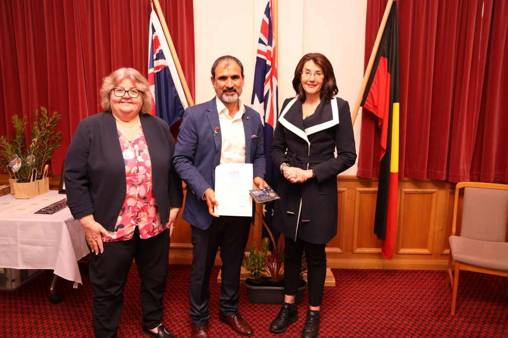 Three people stand indoors in front of red curtains and three flags, including the Australian and Aboriginal flags. The person in the center holds a certificate and booklet. All three are smiling and dressed in business attire.