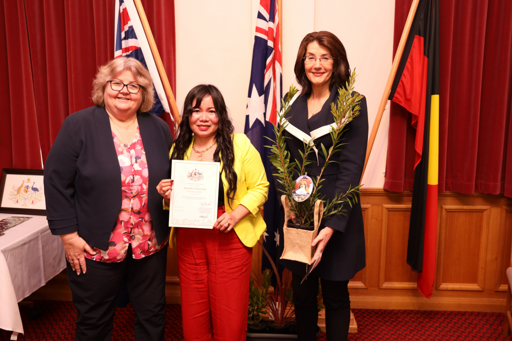 Three women stand indoors in front of Australian and Aboriginal flags. The woman in the center holds a certificate, while the woman on the right holds a potted plant. All are smiling at the camera.
