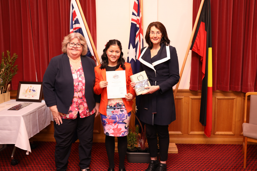 Three women stand indoors in front of Australian and Aboriginal flags. The woman in the center holds a certificate and a small trophy. They are smiling, and a table with framed documents is visible to the left.