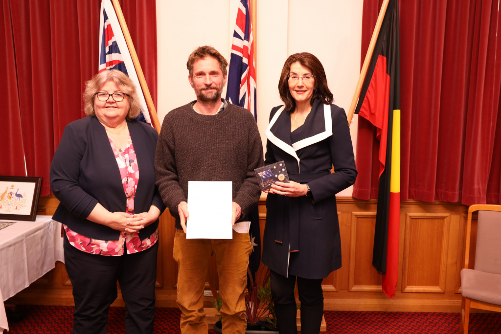 Three people stand indoors in front of red curtains and flags, smiling at the camera. The person in the middle holds a certificate, and the person on the right holds a small box or case.