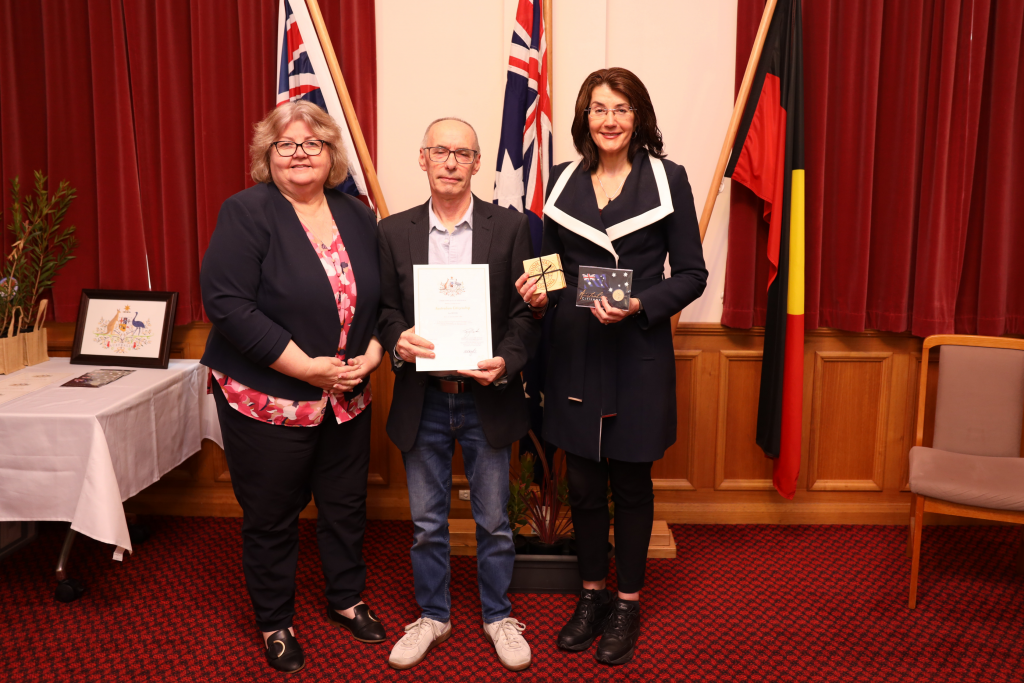 Three people stand indoors in front of Australian and Aboriginal flags. The man in the center holds a certificate; the two women on either side smile. A table with framed items is on the left. The setting appears formal.