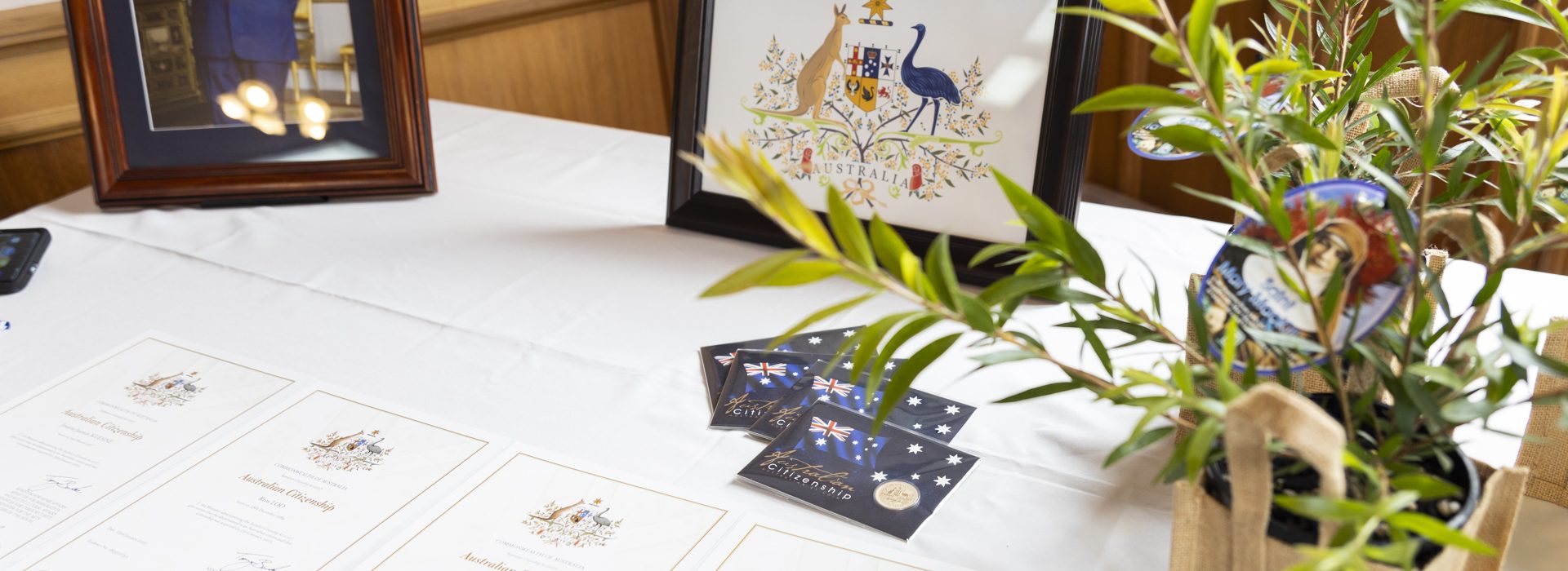 A table displays framed Australian artwork, a framed photo of a man in formal attire, Australian citizenship certificates, postcards, and a small plant in a beige bag.