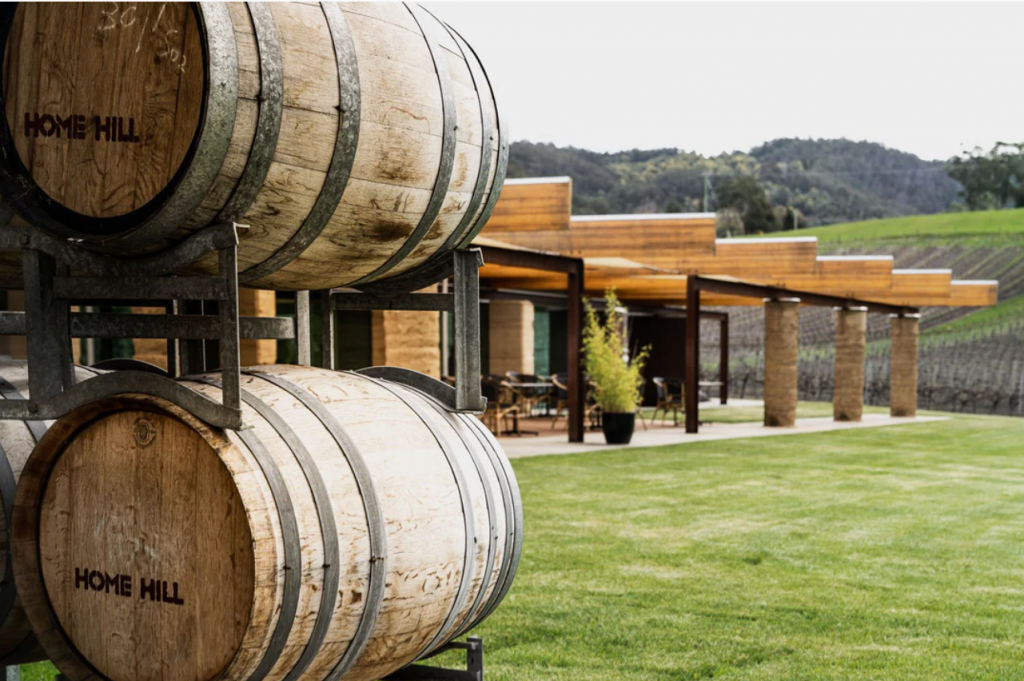 Two large wooden barrels labeled Home Hill are stacked on a metal rack outside a modern winery building, with green grass and vineyard rows in the background.