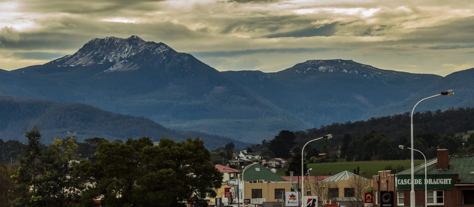 Town buildings with street signs in the foreground, backed by tall, forested mountains under a cloudy sky. The mountain peaks have patches of snow, and trees and lampposts line the streets.