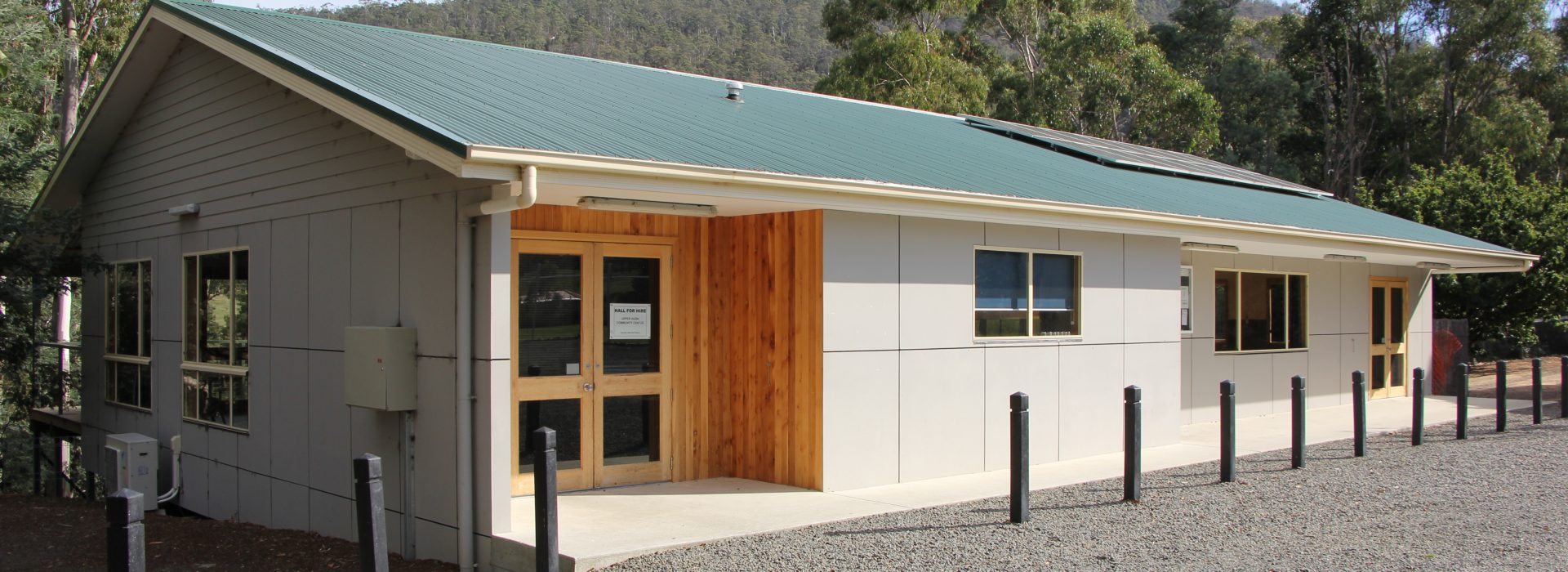 A modern, single-story building with a green roof and beige siding, surrounded by trees and mountains, with a gravel parking area and black bollards in front of the entrance.