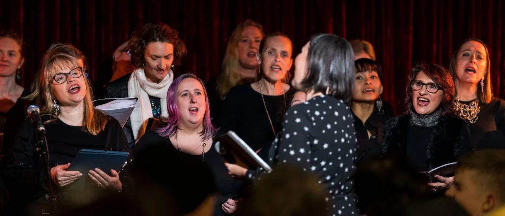A group of women sing together on stage in front of a dark red curtain, holding sheet music, while a conductor stands facing them with her back to the camera.