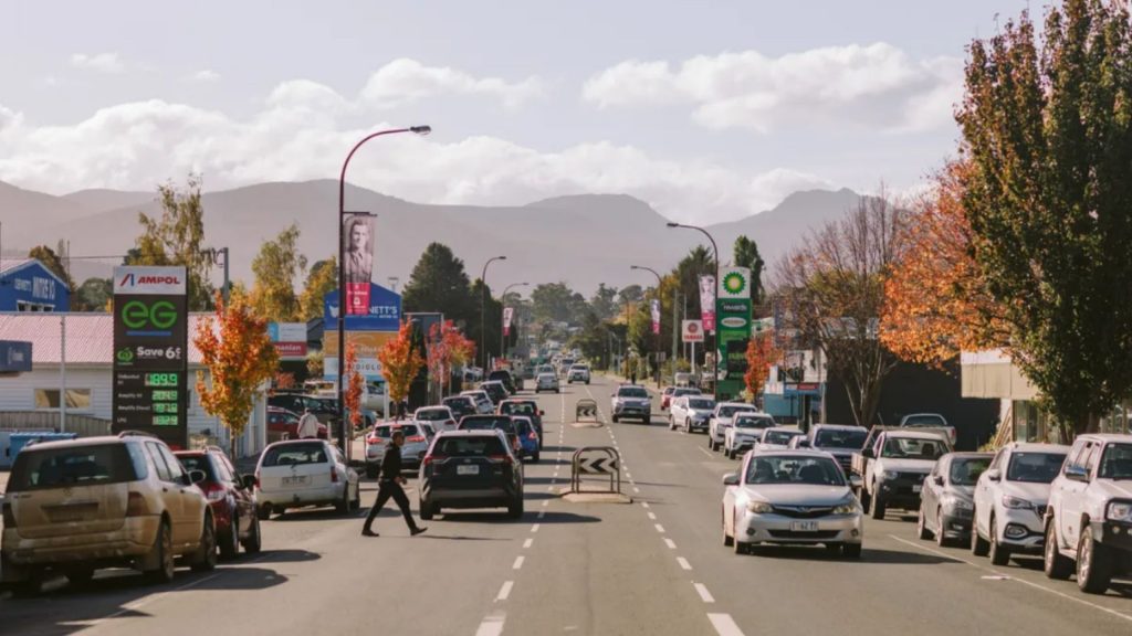 A busy street in a small town with cars parked along both sides, people crossing the road, autumn trees lining the sidewalks, and distant mountains under a partly cloudy sky.