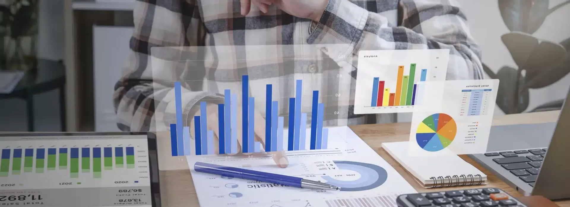 A person in a plaid shirt points at papers with charts on a desk, surrounded by a calculator, notebook, books, and digital bar and pie graphs appearing as holograms above the workspace.