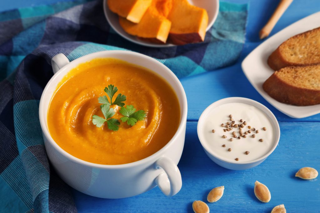 A white bowl of creamy orange pumpkin soup garnished with parsley sits on a blue table, next to a small bowl of sour cream with pepper, toasted bread slices, and pumpkin seeds, with pumpkin pieces in the background.