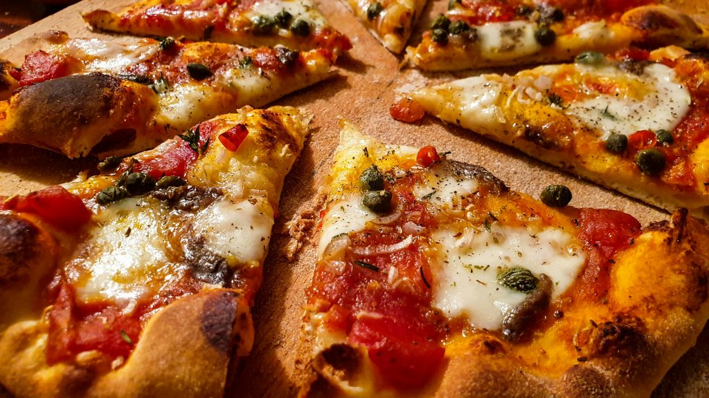 Close-up of several slices of freshly baked pizza with melted cheese, tomato sauce, herbs, capers, and crispy golden-brown crust on a wooden surface.