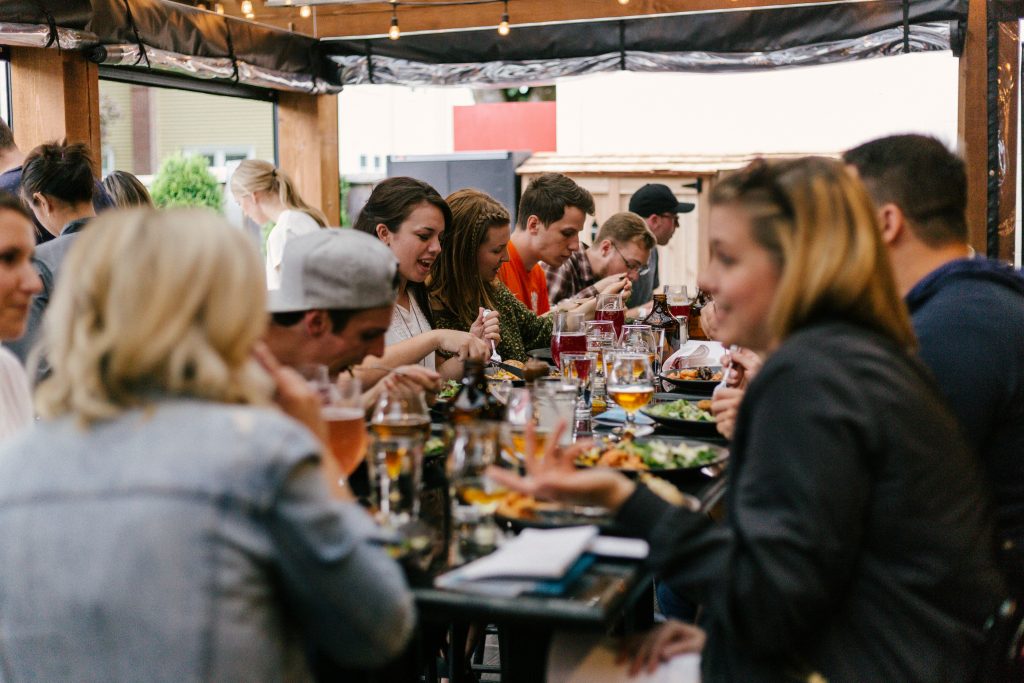 A group of people sitting together at a long outdoor table, enjoying food and drinks, talking, and laughing under string lights in a casual setting.