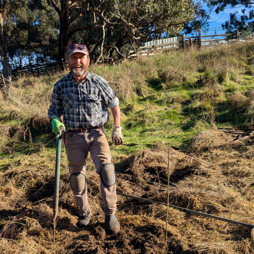 A man in outdoor work clothes, gloves, and knee pads stands on grassy, uneven ground holding a tool, smiling. He is surrounded by dry grass and trees under a clear sky.