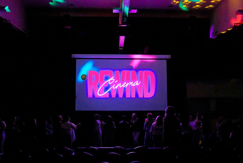 A dark auditorium with people standing in front of a large screen displaying the words REWIND Cinema in bold, colorful letters. Colored lights cast patterns on the ceiling and some audience members.