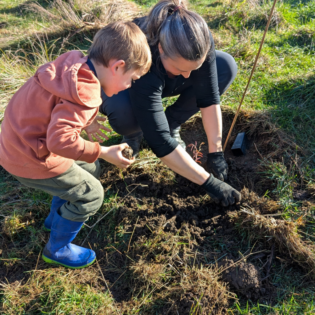 An adult and a child kneel on grass, planting a tree sapling together. The adult wears black gloves and the child wears a brown hoodie and blue boots. They are working in a patch of freshly dug soil.
