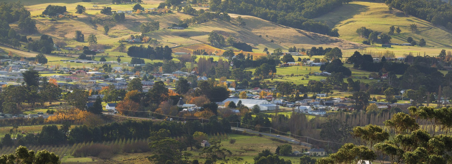 A small rural town stretches across a green valley, surrounded by rolling hills and scattered trees under soft sunlight, with houses, open fields, and forested slopes visible in the landscape.