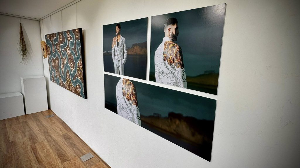 A gallery wall displays a large abstract painting with earthy tones and three portrait photos of a man in a patterned suit with tattooed arms, standing against a dark, blurred landscape background. Dried plants hang on the wall nearby.