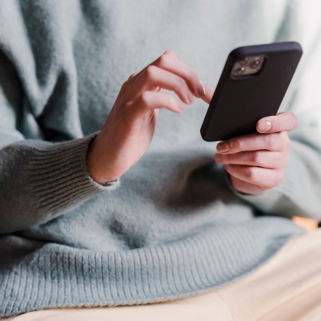 A person wearing a light blue sweater is holding and using a smartphone, with their focus on the device. Only their hands and torso are visible in the image.