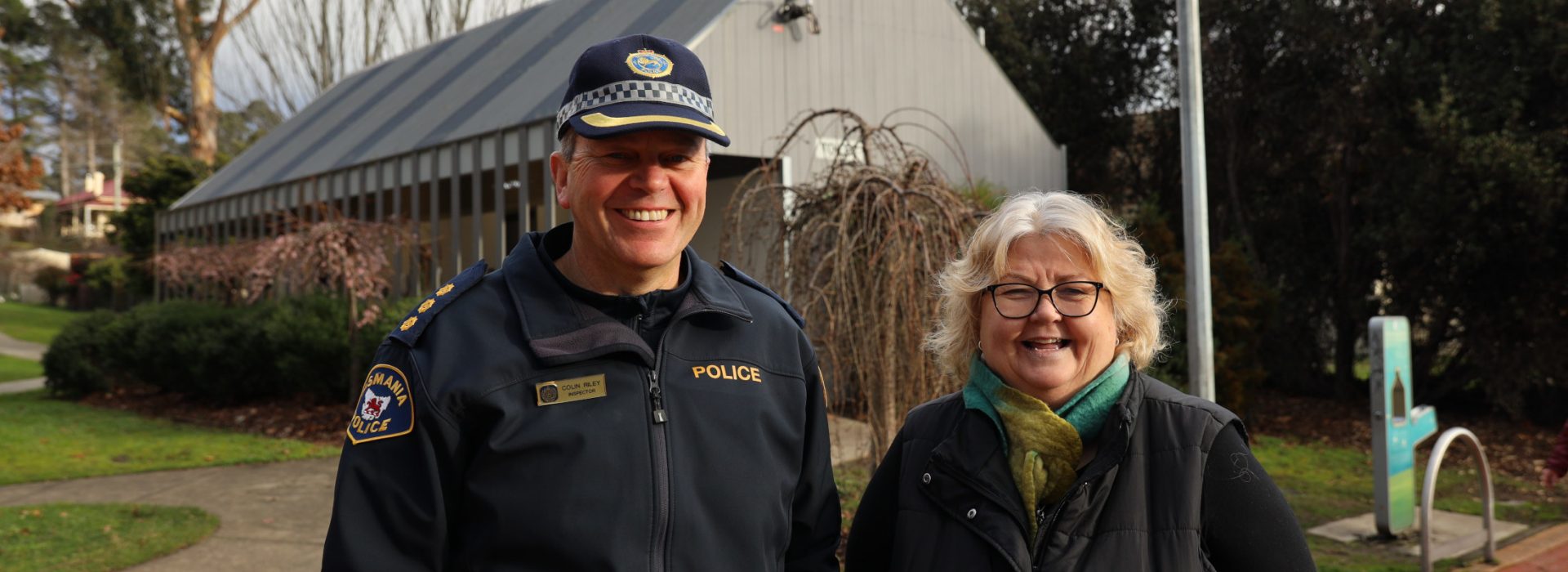 A smiling police officer and a woman stand outside on a paved path, with a modern building, trees, and bushes in the background. The officer wears a uniform and cap, while the woman wears glasses, a scarf, and a vest.