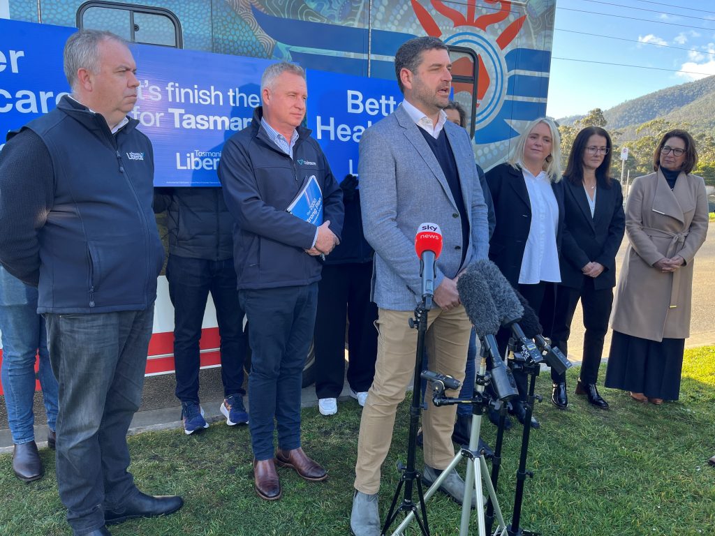 A group of people, including several men and women, stand outdoors near microphones and a Liberal campaign sign as one man speaks to the media. Hills and trees are visible in the background.