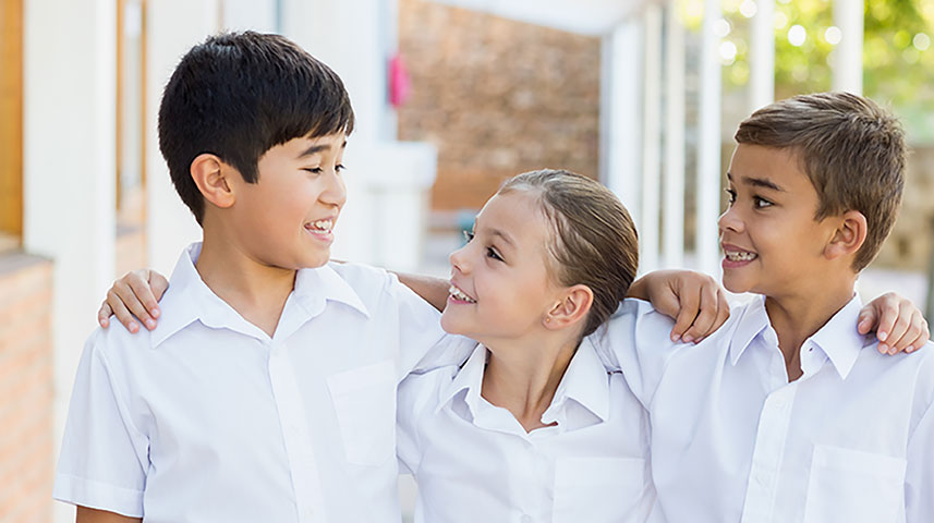 Three children in white school uniforms smile and walk together outdoors, with their arms around each other’s shoulders, showing friendship and happiness.