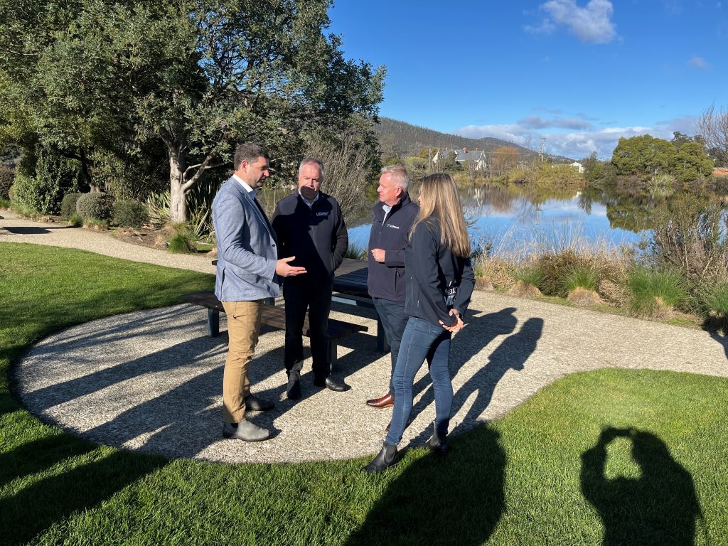 Four people stand talking on a sunny path beside a lake and grassy area, with trees and hills in the background. A fifth person’s shadow is visible in the foreground, holding up a camera to take the photo.