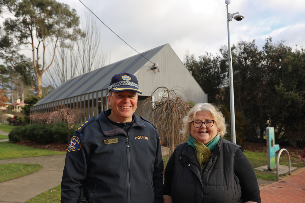A smiling police officer and a woman stand outdoors in front of a modern building with a sloped roof, surrounded by trees and greenery on a cloudy day.