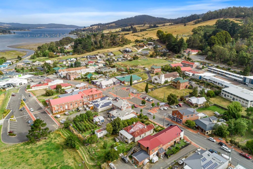 Aerial view of a small town with red-roofed buildings, green fields, trees, winding roads, and a river with moored boats in the background, under a blue sky with light clouds.
