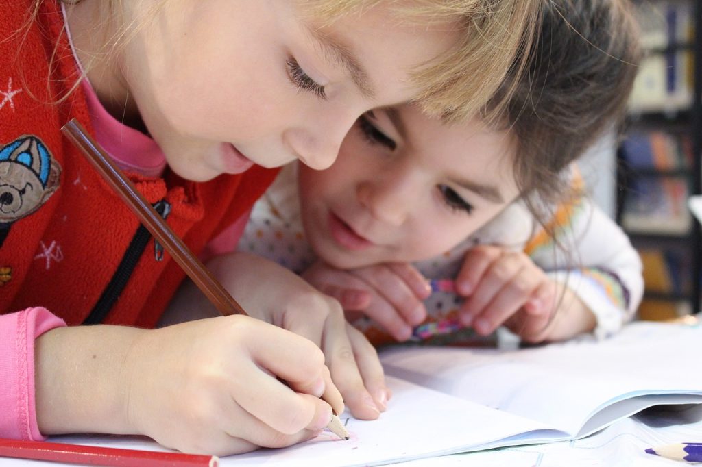 Two young children are closely drawing or writing in a notebook together, one holding a pencil while the other watches attentively. They appear engaged and focused on their activity.