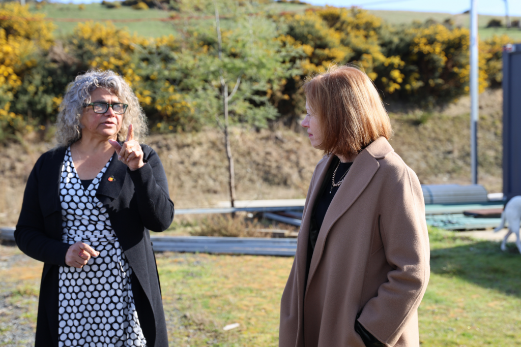 Two women stand outdoors, engaged in conversation. One woman gestures with her hand while the other listens. Both wear coats; the setting is grassy with some construction materials and shrubbery in the background.
