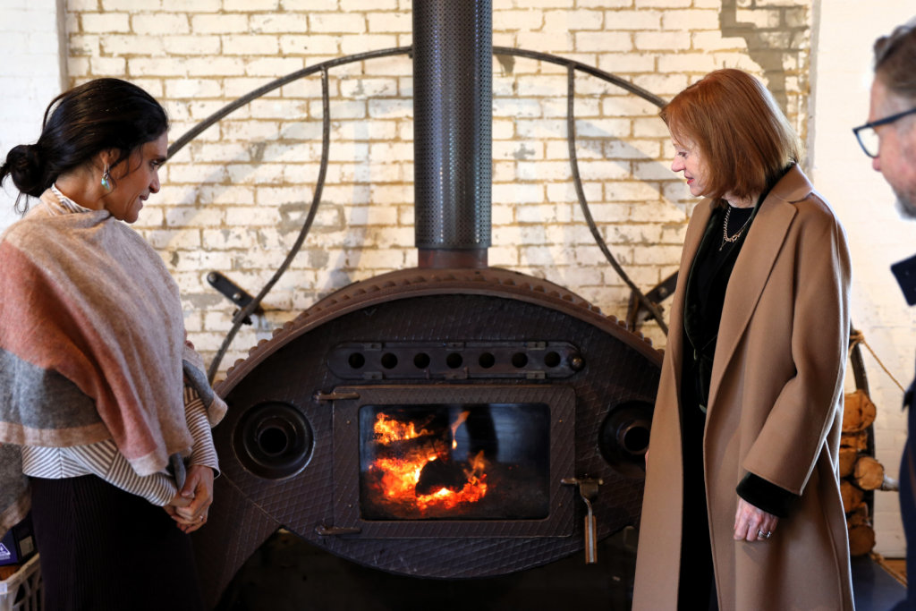 Two women stand facing a lit wood-burning stove with a glass door, while a man stands partially visible on the right. The stove is in front of a white brick wall. The women are wearing winter coats.