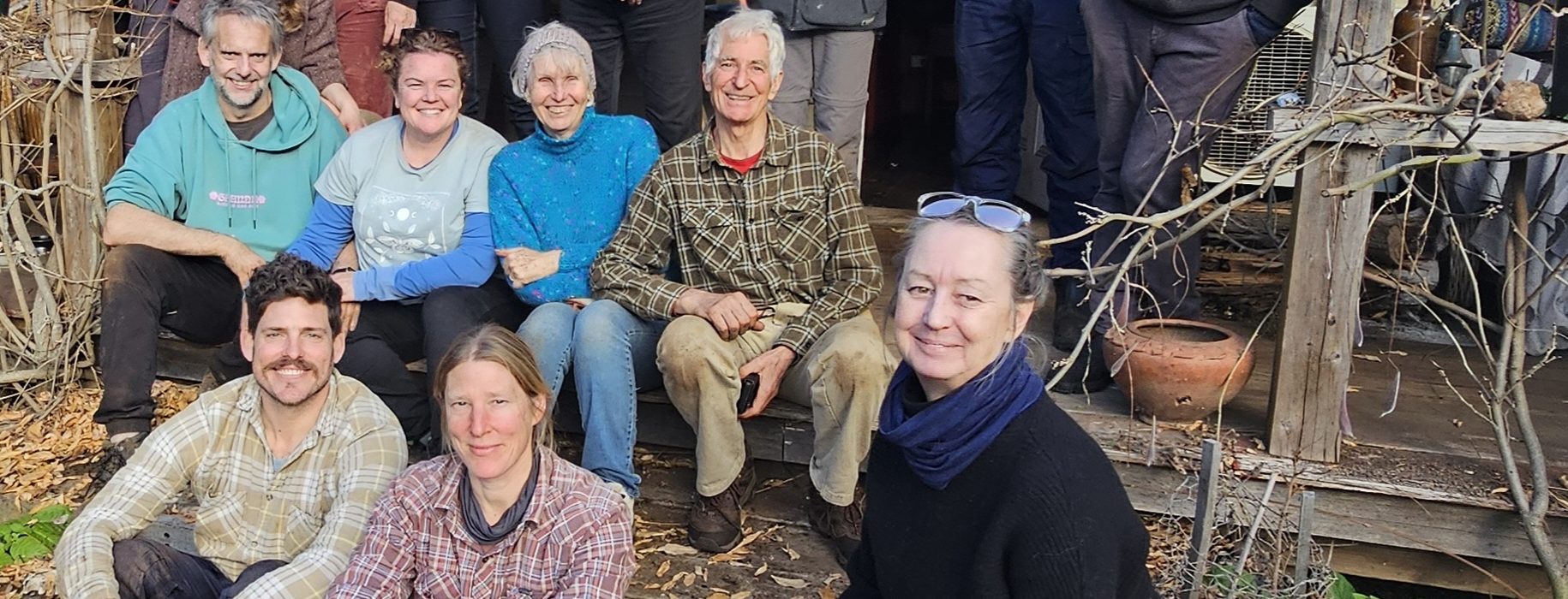A group of people of varying ages pose and smile together on the porch and steps of a rustic wooden house, surrounded by plants and garden pots on a cloudy day.