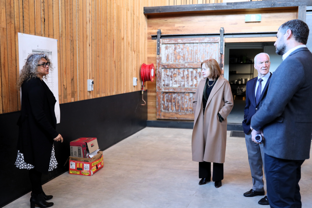 Four people stand in a large room with wooden walls; one woman gestures toward a wall with papers, while the other three—two men and a woman—listen attentively near a rustic wooden door. Boxes and a fire hose are visible.