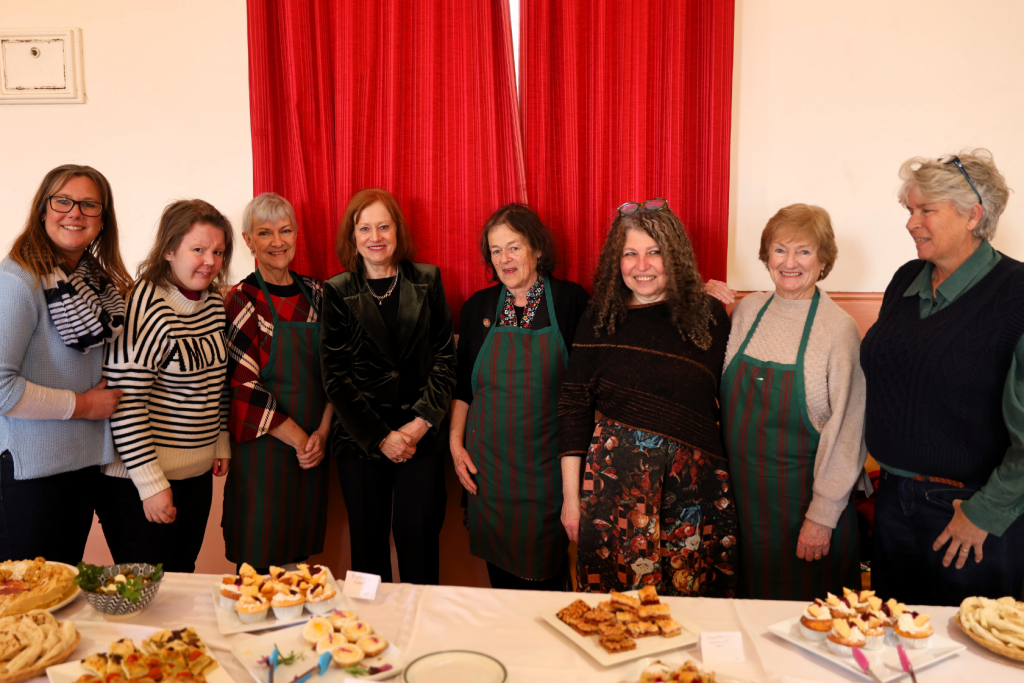 Eight women, some wearing green-striped aprons, stand together and smile behind a table filled with various pastries and finger foods. They are in a room with red curtains and cream-colored walls.