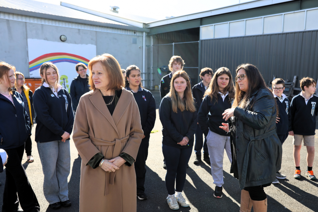 A group of students and two adult women stand outside a school building. One woman in a tan coat listens while the other in a dark jacket talks. A rainbow mural is visible in the background.