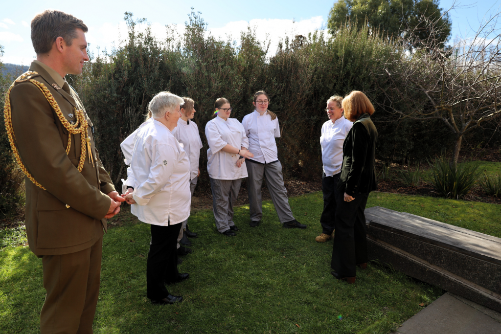 A group of six people in chef uniforms and one person in a military uniform stand outdoors on grass, talking to a woman in a black jacket near a stone bench with bushes in the background.